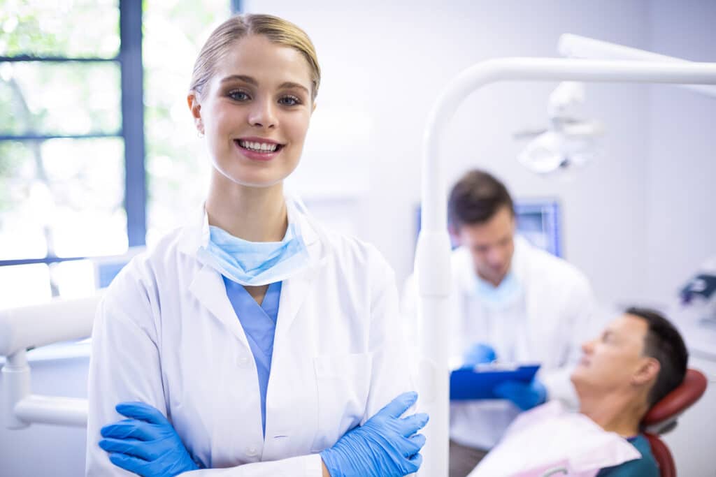 Smiling female dentist standing with arms crossed in a modern dental clinic, while a male dentist attends to a patient in the background